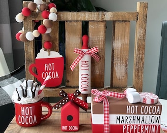 May include: A tiered wooden shelf display with a red and white gingham ribbon and a red and white bead garland. The shelf is decorated with a red and white hot cocoa themed decor, including a rolling pin, a mug, a birdhouse, and a stack of books with the words "Hot Cocoa, Marshmallow, Peppermint".