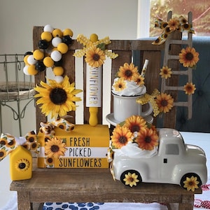 May include: A decorative display featuring a rustic wooden backdrop with various sunflower-themed items. Includes a white ceramic truck filled with sunflowers, a mug with a whipped cream topping and sunflowers, and a small wooden ladder adorned with sunflowers and bows. The display also has a wooden sign that says "FRESH PICKED SUNFLOWERS".