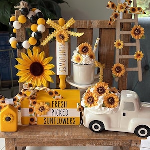 May include: A rustic display featuring a wooden ladder, a white ceramic truck, and a mug, all adorned with sunflowers. A wooden sign reads "FRESH PICKED SUNFLOWERS". Decorative beads and a rolling pin with "SUNFLOWER" text complete the arrangement.