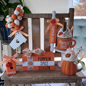 May include: A miniature wooden bench with fall-themed decorations. There is a pumpkin spice rolling pin, a pumpkin spice mug, a pumpkin spice season block, a small birdhouse, and a white sign with a heart cutout.