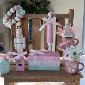 May include: A pink and white themed Christmas display with a nutcracker, a rolling pin with the words "Sweet Shop" on it, a pink mug with the words "Sugar Plum" on it, and a stack of three boxes with the words "Ballet Nutcracker Sugar Plums" on them.