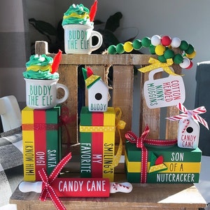 May include: A set of Christmas-themed wooden blocks with various sayings, including "Buddy the Elf", "My Favorite", "Smiling's", "Cotton Headed Ninny Muggins", "Son of a Nutcracker", and "Candy Cane". The blocks are stacked on top of each other and decorated with red and green ribbon. There is also a rolling pin with the words "Candy Cane" written on it.