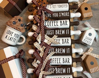 May include: A collection of decorative items including miniature rolling pins with text like 'COFFEE BAR' and 'JUST BREW IT', small mugs labeled 'LOVE YOU A LATTE', wooden blocks reading 'MORE COFFEE PLEASE', and house-shaped signs labeled 'COFFEE' or 'SUGAR PLEASE'.