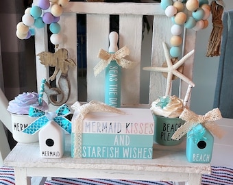 May include: A white wooden bench with a blue and white striped fabric on the bottom shelf.  The bench is decorated with a variety of nautical themed items, including a mermaid figurine, a starfish, a seashell, and a wooden sign that reads "Mermaid Kisses and Starfish Wishes".