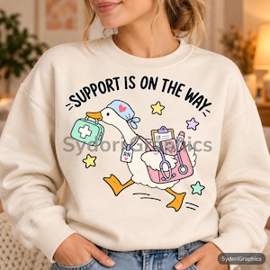 May include: A cream-coloured sweatshirt featuring a graphic of a white goose wearing a blue nurse cap, carrying a light green medical bag and a pink medical tote containing a clipboard, stethoscope, and a notebook labelled RN NOTES.