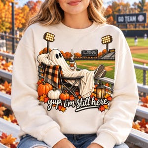 May include: A cream-colored sweatshirt featuring a graphic of a ghost sitting in a folding chair at a baseball field, holding a coffee cup. The design includes pumpkins, a lantern, autumn leaves, and the text "yup, i'm still here" in black script.