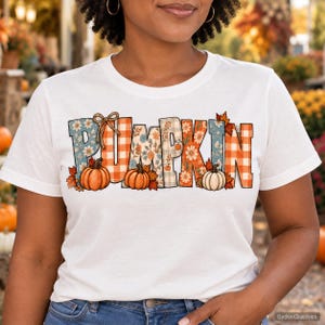 May include: A white short-sleeve t-shirt featuring the word PUMPKIN in large block letters. Each letter displays a unique pattern including orange and white gingham, blue floral prints, and autumn leaves. Three orange pumpkins are at the base.