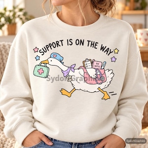 May include: A cream-coloured crewneck sweatshirt featuring a graphic of a white goose dressed as a nurse, carrying a medical kit and bag. The text reads SUPPORT IS ON THE WAY, accompanied by a clipboard, RN NOTES notebook, stethoscope, and colourful stars.