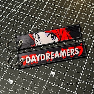 May include: Two black and red fabric keychains. The top keychain features a cartoon woman with red hair and blue eyes. The bottom keychain has the word "DAYDREAMERS" in white letters.