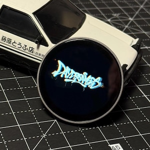 May include: A round, black pop socket with the word "Daydreamers" in a neon blue graffiti style. The pop socket has a silver rim. A white toy car is in the background.