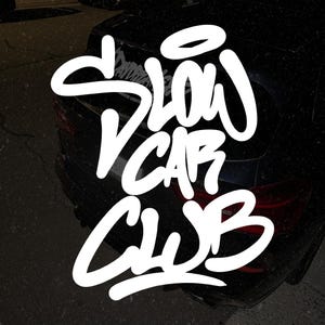 May include: White "SLOW CAR CLUB" text in a graffiti style over the rear of a black car. The text is large and stylized, with a slight shadow effect. The car's taillights are visible.