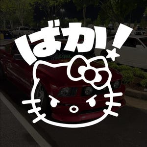May include: A white graphic featuring Japanese characters and a Hello Kitty face overlayed on a red sports car. The car is parked in a lot with other vehicles. The Japanese text says "Baka!"