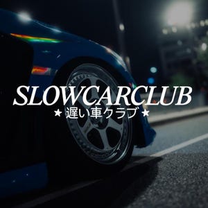 May include: A blue car wheel and fender at night with the white text SLOWCARCLUB and the Japanese characters 遅い車クラブ with stars on either side. The metallic wheel features a multi-spoke design with a polished rim edge and visible lug nuts.