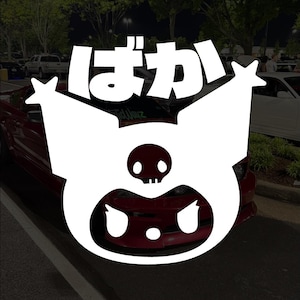 Puede incluir: Calcomanía de vinilo blanco con un personaje de dibujos animados estilizado con una cabeza en forma de calavera y una gran boca abierta. Los caracteres japoneses "ばか" están encima del personaje.