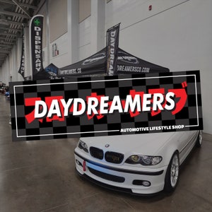 May include: A modified white car is parked in front of a banner displaying "DAYDREAMERS AUTOMOTIVE LIFESTYLE SHOP." The banner features a black and white chequered pattern and red Japanese characters.