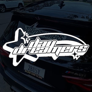 May include: White vinyl decal featuring the word "daydreamers" in a stylized font, with a star and diamond design. The decal is applied to the rear window of a black car. The license plate reads "2BUSY".