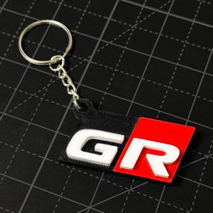 以下が含まれることがあります： 黒と赤の背景に白い文字「GR」が配置されたキーホルダー。 キーホルダーは、シルバーの金属リングとチェーンに取り付けられています。 大胆でグラフィカルなデザインです。