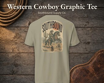 May include: A light olive green t-shirt featuring a graphic on the back. The design depicts a cowboy riding a horse in a desert landscape with a cactus, the text SOUTHBOUND, and the initials SBC. The shirt has short sleeves and a crew neck.