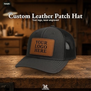 Puede incluir: Una gorra tipo camionero de color gris carbón con un parche rectangular de cuero color canela en la parte frontal. El parche tiene grabado con láser el texto YOUR LOGO HERE en una fuente oscura. La gorra tiene un panel frontal rígido.