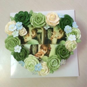 May include: A heart-shaped cake decorated with green, white, and blue buttercream flowers. The cake has a photo collage of two people on the front.