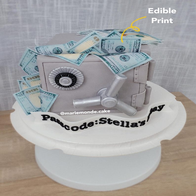 May include: A gray safe cake decorated with edible dollar bills. The safe has a silver handle and a round dial. The cake is on a white cake stand with the words "Passcode: Stella's Pay" written in black icing.