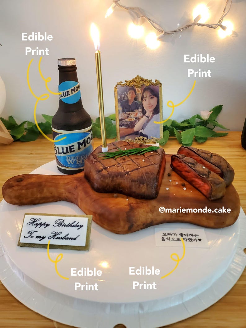 May include: A birthday cake decorated with a chocolate steak, a bottle of beer, and a photo of a couple. The cake is on a white plate and has a sign that says "Happy Birthday to my Husband".