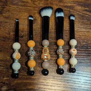 May include: Five make-up brushes with black handles and decorative beaded bases. The beads are in shades of orange, cream, brown, and black, with some featuring floral and animal designs. The brush heads are white and black.