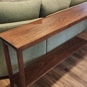 May include: A long, narrow console table crafted from dark brown wood with a prominent, swirling grain pattern. The table features a smooth, polished rectangular top surface and a lower shelf supported by four slender, straight wooden legs.