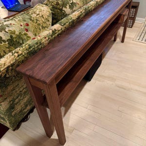 May include: A long, dark brown wooden console table with a lower shelf. The table has a rectangular top and angled legs. The wood grain is visible. The table is placed against a floral patterned sofa.