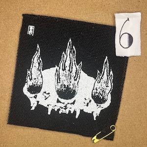 May include: Black square patch featuring a white graphic of flames and a skull. A small piece of paper with the number "6" is next to the patch. A gold safety pin is also in the image.