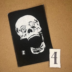 May include: A black fabric patch featuring a white, screaming skull design. The patch is rectangular with a stitched border. A small, white number "4" is on a separate, small, rectangular piece of paper.