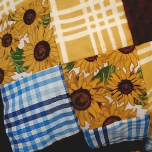 May include: A patchwork quilt featuring squares of fabric with various patterns. Some squares display sunflowers, others have yellow and white plaid, and some are blue and white plaid. The quilt also has brown squares.