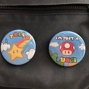 May include: Two blue and white button pins with a video game theme. The first pin features a yellow star with a rainbow and the text "TRIED". The second pin features a red mushroom with the text "I'M JUST A FUNGI".