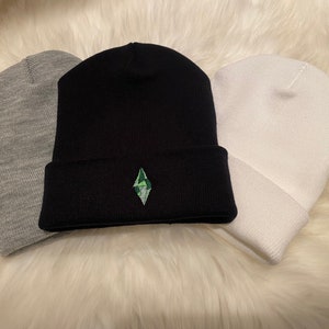 Può includere: Tre berretti di lana in grigio, nero e bianco. Il berretto nero ha un logo a forma di diamante verde e nero ricamato sul davanti.