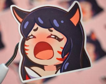 May include: A sticker featuring a cartoon character with black hair, red markings on her face, and a surprised expression. She is wearing a white shirt and has cat ears.
