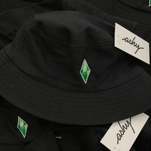 Può includere: Cappello a secchiello nero con un logo a forma di diamante verde ricamato sul davanti. Il cappello ha un'ampia tesa e una corona piatta. Il logo è un diamante stilizzato con un contorno verde e un interno bianco.