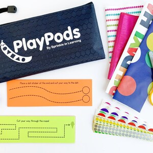 May include: A black zippered pouch with the text "PlayPods By Sprinkle in Learning" and a green peapod logo. The pouch contains a set of colorful craft supplies, including scissors, patterned paper, and sticker sheets. The supplies are for a craft activity that involves cutting and sticking.