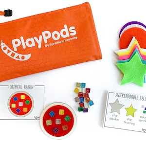 May include: A set of felt food play pieces for kids, including a green star, a yellow star, a red circle, a purple circle, and a white circle. The set also includes two recipe cards, one for oatmeal raisin cookies and one for snickerdoodle cookies. The cards have pictures of the cookies and list the ingredients, including star cookie, star frosting, and colored sprinkles.