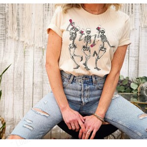 Puede incluir: Una camiseta beige claro con un gr&aacute;fico de tres esqueletos bailando y sosteniendo copas de vino. Los esqueletos est&aacute;n en blanco y negro.