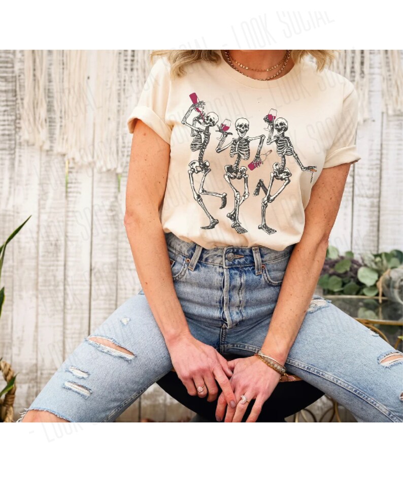 Puede incluir: Una camiseta beige claro con un gr&aacute;fico de tres esqueletos bailando y sosteniendo copas de vino. Los esqueletos est&aacute;n en blanco y negro.