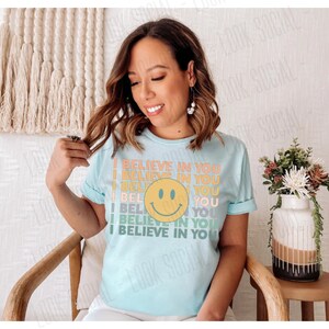 May include: A light blue t-shirt with the text "I BELIEVE IN YOU" in a retro font and a yellow smiley face. The shirt is short-sleeved and has a crew neck. The design is in shades of orange, yellow, and green.