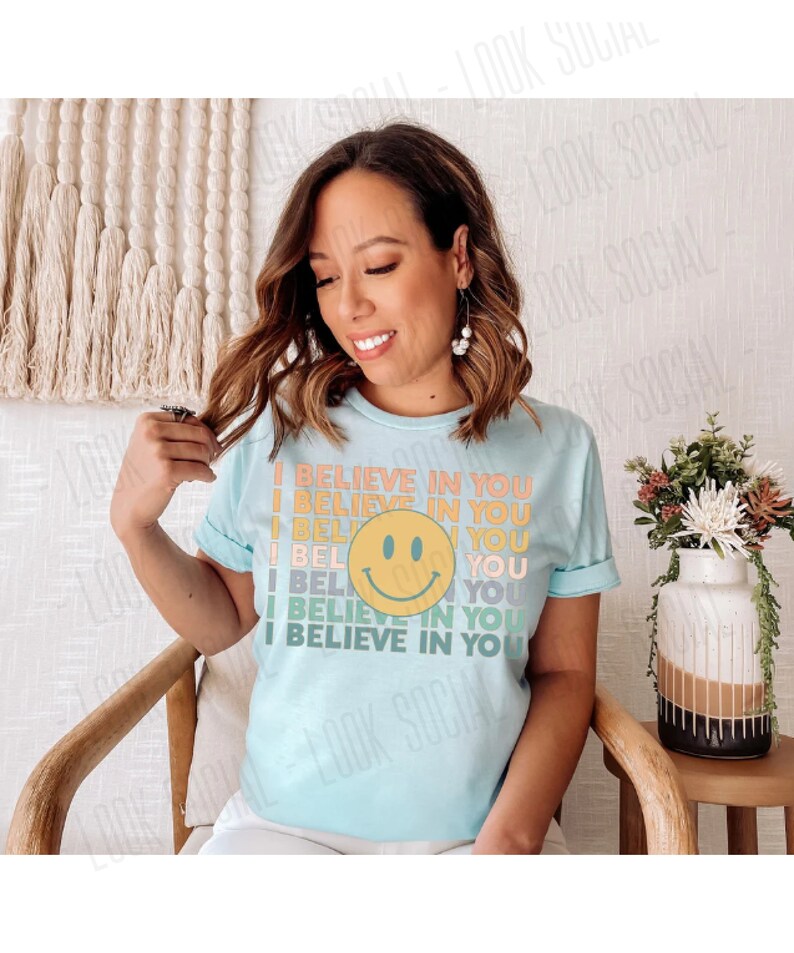 May include: A light blue t-shirt with the text "I BELIEVE IN YOU" in a retro font and a yellow smiley face. The shirt is short-sleeved and has a crew neck. The design is in shades of orange, yellow, and green.