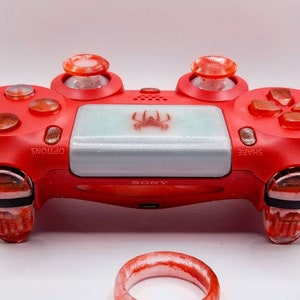 May include: Red PlayStation 4 controller with custom orange and clear thumbsticks and buttons. The center pad is light blue with a spider graphic. A matching ring is in the foreground.
