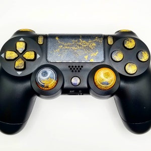 May include: Black PlayStation 4 controller with gold accents on the buttons and touchpad. The directional pad and action buttons are gold. The touchpad has a gold splatter design. The thumbsticks are gold and orange.