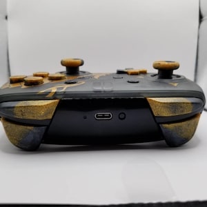 May include: A black Nintendo Switch controller with gold accents and custom buttons. The controller has a textured finish and a USB-C port on the bottom.