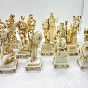 以下が含まれることがあります： 金色のアクセントが付いた白いギリシャ神と女神の置物10個セット。各置物は、金色の文字で神または女神の名前が書かれた台座の上に置かれています。置物は白い表面に一列に並べて配置されています。