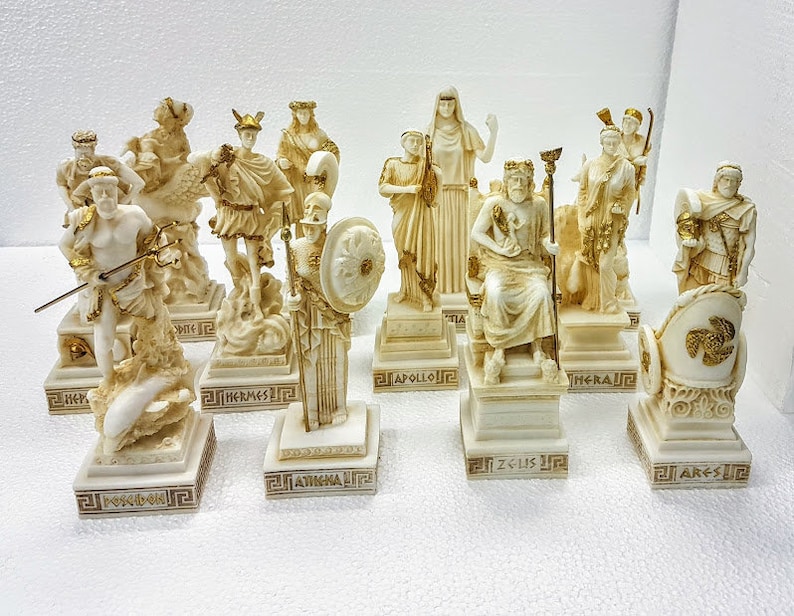 以下が含まれることがあります： 金色のアクセントが付いた白いギリシャ神と女神の置物10個セット。各置物は、金色の文字で神または女神の名前が書かれた台座の上に置かれています。置物は白い表面に一列に並べて配置されています。