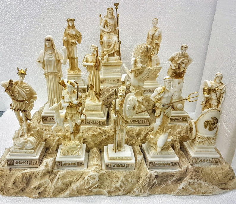 以下が含まれることがあります： ギリシャの神々や女神を表現した、白い樹脂製の置物12個のセット。各置物は、神や女神の名前が刻まれた小さな台座の上に立っています。置物は、茶色の台座の上に半円形に配置されています。