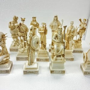 以下が含まれることがあります： 白い大理石製のギリシャ神々や女神の置物12個セット。各置物は、神や女神の名前が金文字で刻まれた正方形の台座の上に立っています。置物はすべて似たようなスタイルで、細部まで丁寧に作られており、流れるようなローブを着ています。