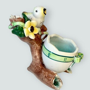 May include: A ceramic bird-shaped planter. The planter has a white body and a yellow beak, perched on a brown branch with green leaves and a yellow flower. The planter has a white egg-shaped container with green stripes.